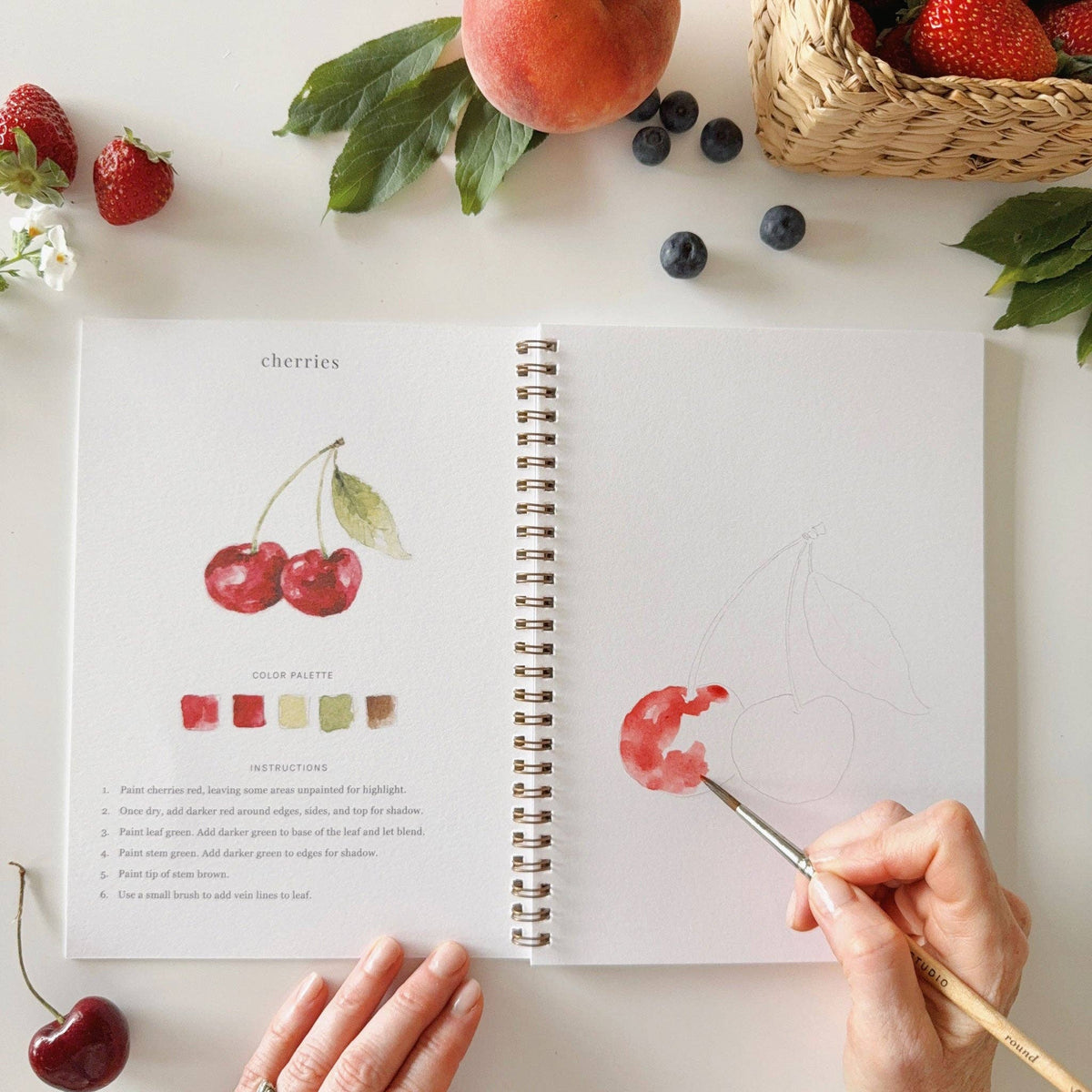 Open spread of Emily Lex Studio Fruit Watercolor Workbook at the cherries lesson — completed red cherries illustration with color palette and 6-step instructions on the left, hand beginning to paint red watercolor on the outlined cherries sketch on the right, surrounded by fresh fruit props