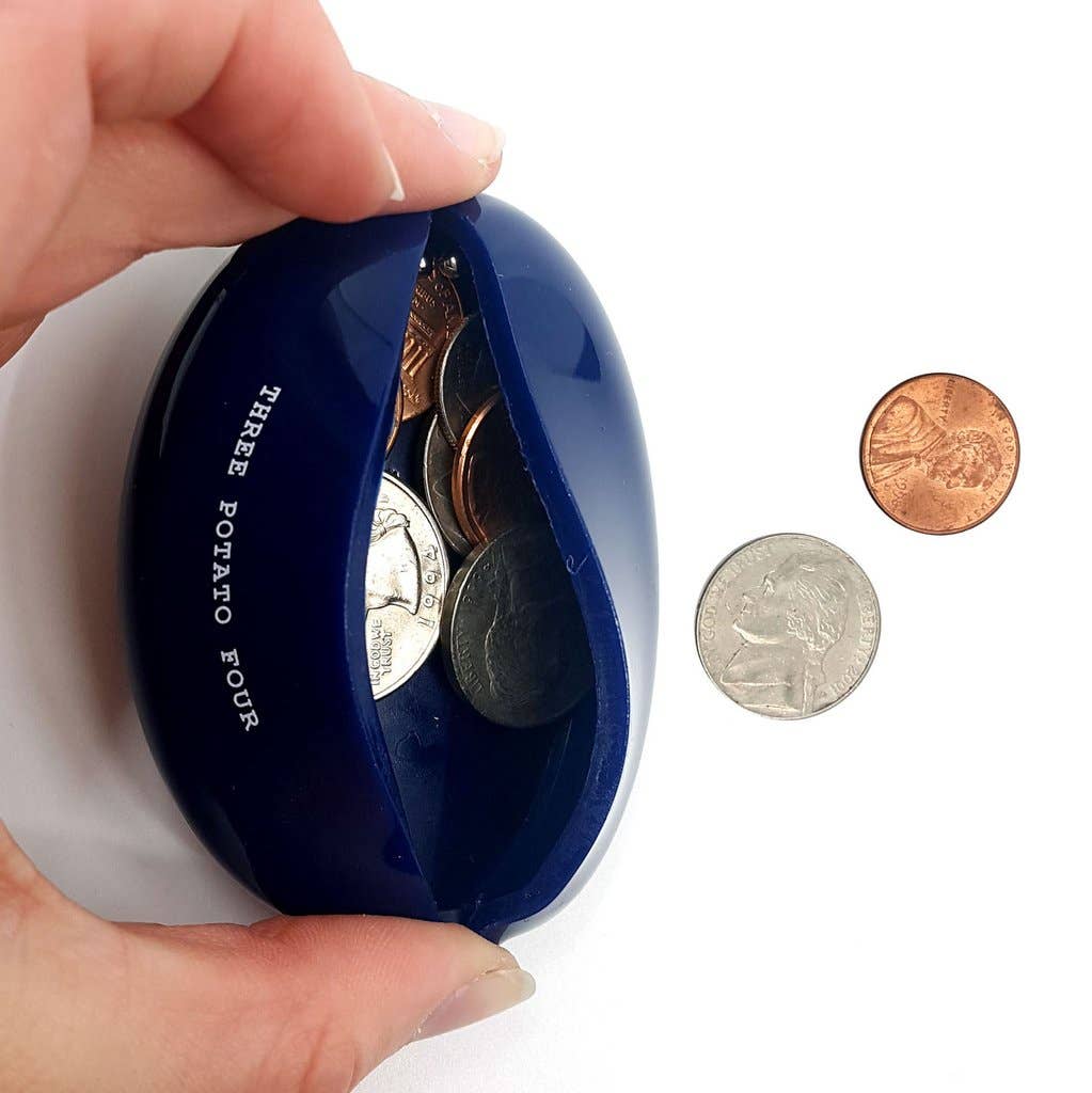 Hand squeezing open the Three Potato Four Change Is Good rubber coin pouch on white background — cobalt blue oval pouch squeezed open at top seam revealing a mix of US coins inside including quarters and pennies; "THREE POTATO FOUR" text visible in yellow on the back face; a dime and a penny shown separately to the right for scale