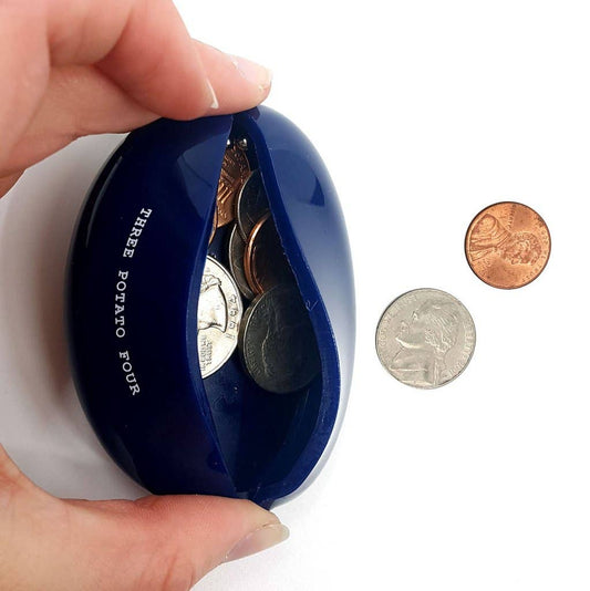 Hand squeezing open the Three Potato Four Change Is Good rubber coin pouch on white background — cobalt blue oval pouch squeezed open at top seam revealing a mix of US coins inside including quarters and pennies; "THREE POTATO FOUR" text visible in yellow on the back face; a dime and a penny shown separately to the right for scale
