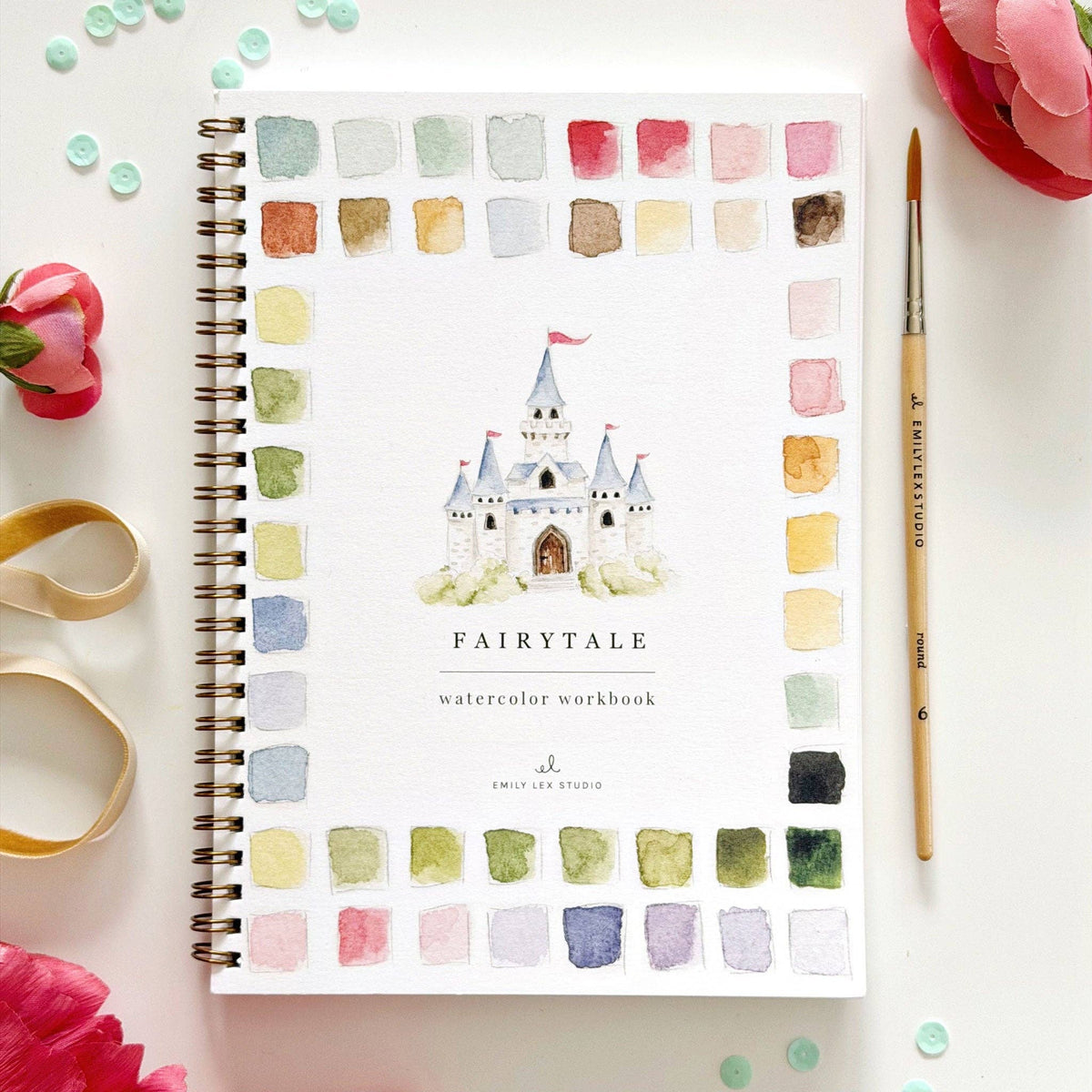 Fairytale Watercolor Workbook by Emily Lex Studio — cover flat lay on white surface with pink ranunculus flowers, gold ribbon, teal sequins, and Emily Lex round size 6 paintbrush at right; cover features a watercolor castle with pink flag turrets, "FAIRYTALE watercolor workbook" text, Emily Lex Studio logo, and a full border of soft pastel color swatches in pinks, mints, sage greens, lavenders, and taupes; gold spiral binding on left