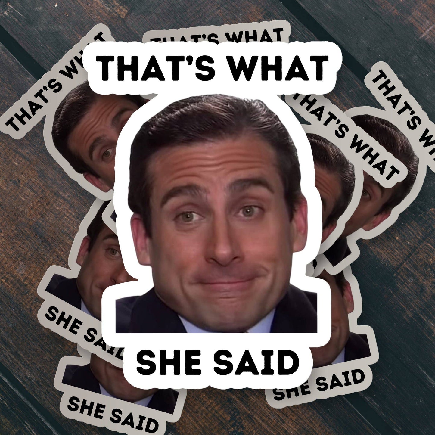 Stack of Sticker Bull That's What She Said die cut vinyl stickers spread in a radial fan on a dark wood surface — photo-real Michael Scott smiling with bold black "THAT'S WHAT / SHE SAID" text; scalloped white die cut borders