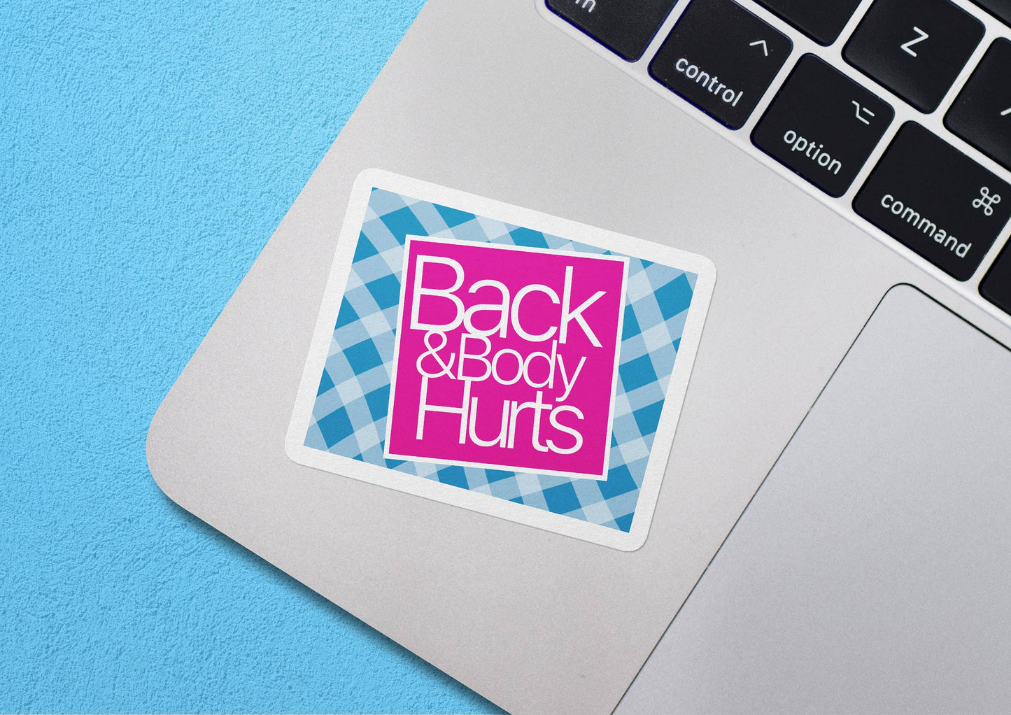 Sticker Bull Back & Body Hurts vinyl sticker applied to a MacBook trackpad against a light blue background — hot pink square with bold white script over blue gingham