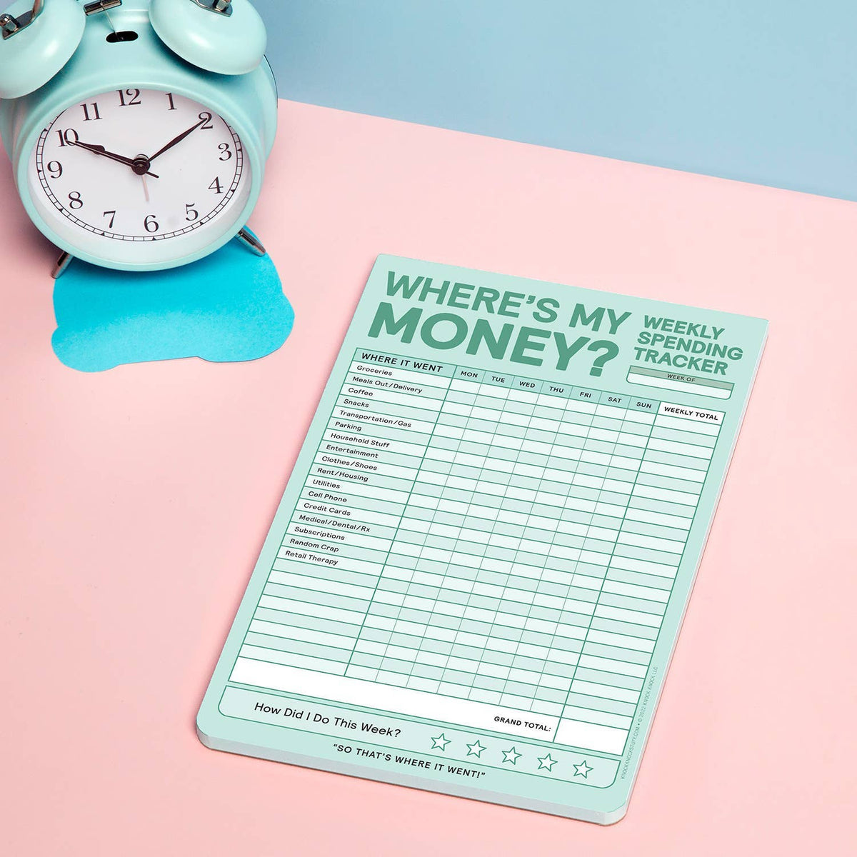 Where's My Money? Weekly Spending Tracker notepad lying flat on a pink surface beside a mint blue retro alarm clock — showing full grid layout on a split blue and pink background