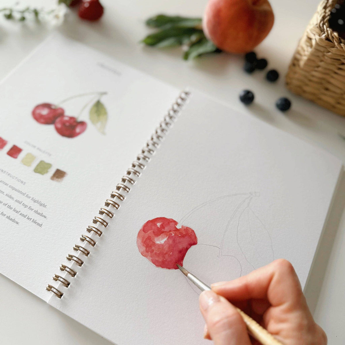Close-up of hand painting the cherries lesson in the Emily Lex Studio Fruit Watercolor Workbook, brush adding red watercolor to the first cherry on the sketch page beside the completed reference illustration, with fresh blueberries and peach in the background