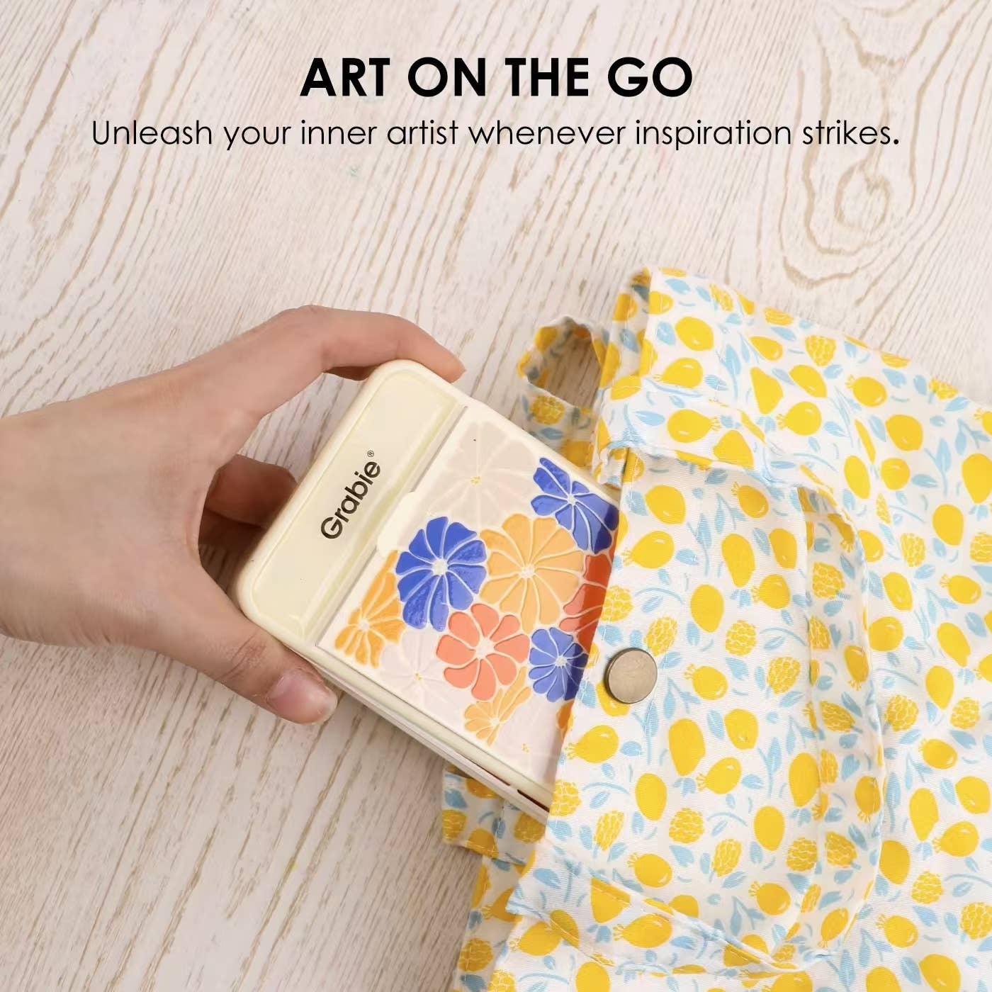Hand holding the compact closed Grabie watercolor set with floral print cover being slipped into a jacket pocket, with "Art on the Go" text overlay