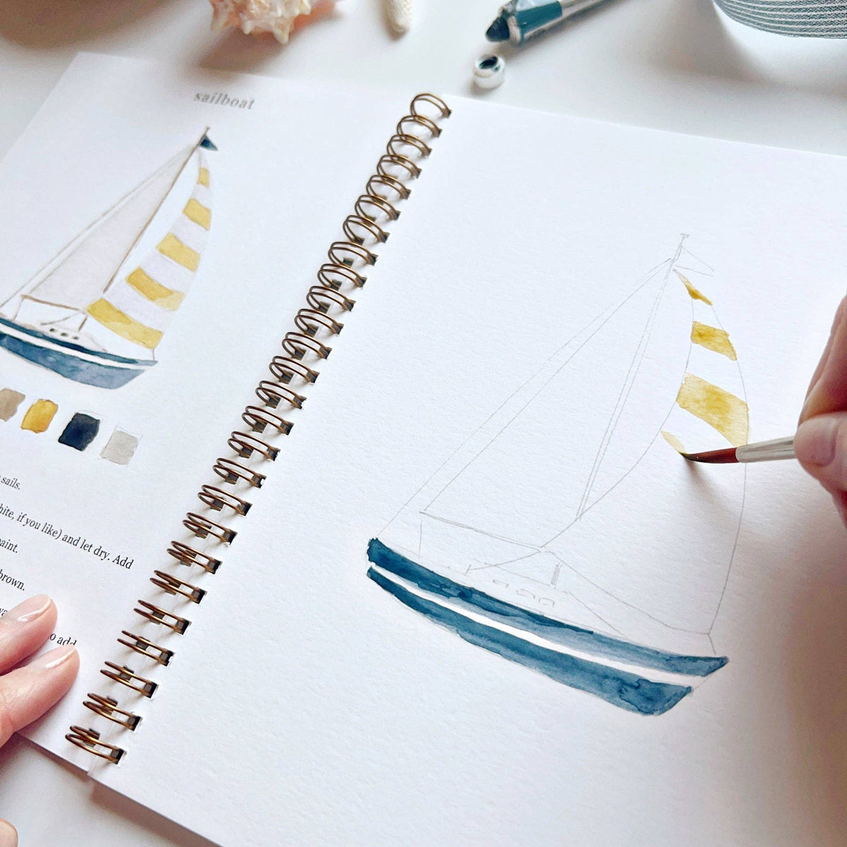 Angled close-up of Seaside Watercolor Workbook open to sailboat page, hand painting yellow stripes on the sails and blue hull of a partially painted sailboat sketch, with completed illustration and color palette partially visible on left