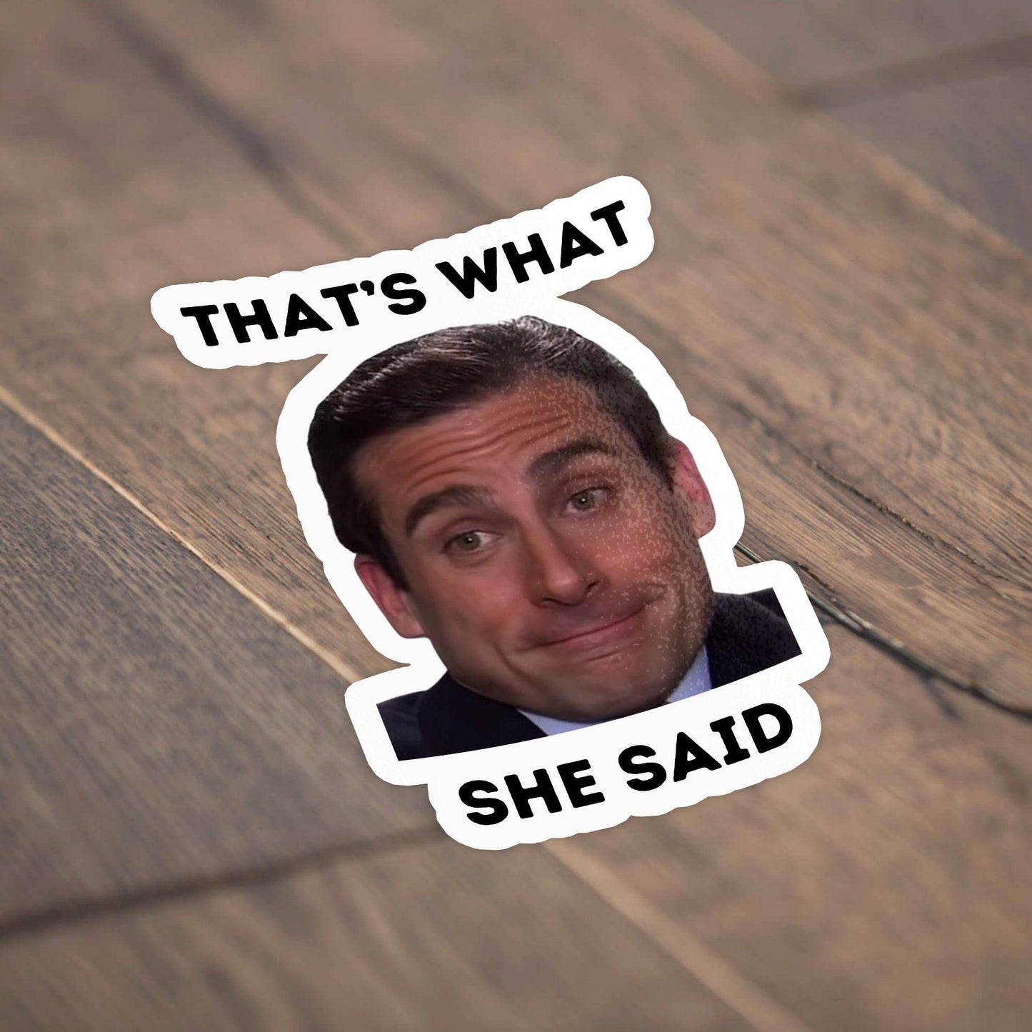 Sticker Bull That's What She Said die cut vinyl sticker lying flat on a light diagonal wood surface — photo-real Michael Scott smiling with bold black "THAT'S WHAT / SHE SAID" text; scalloped white die cut border