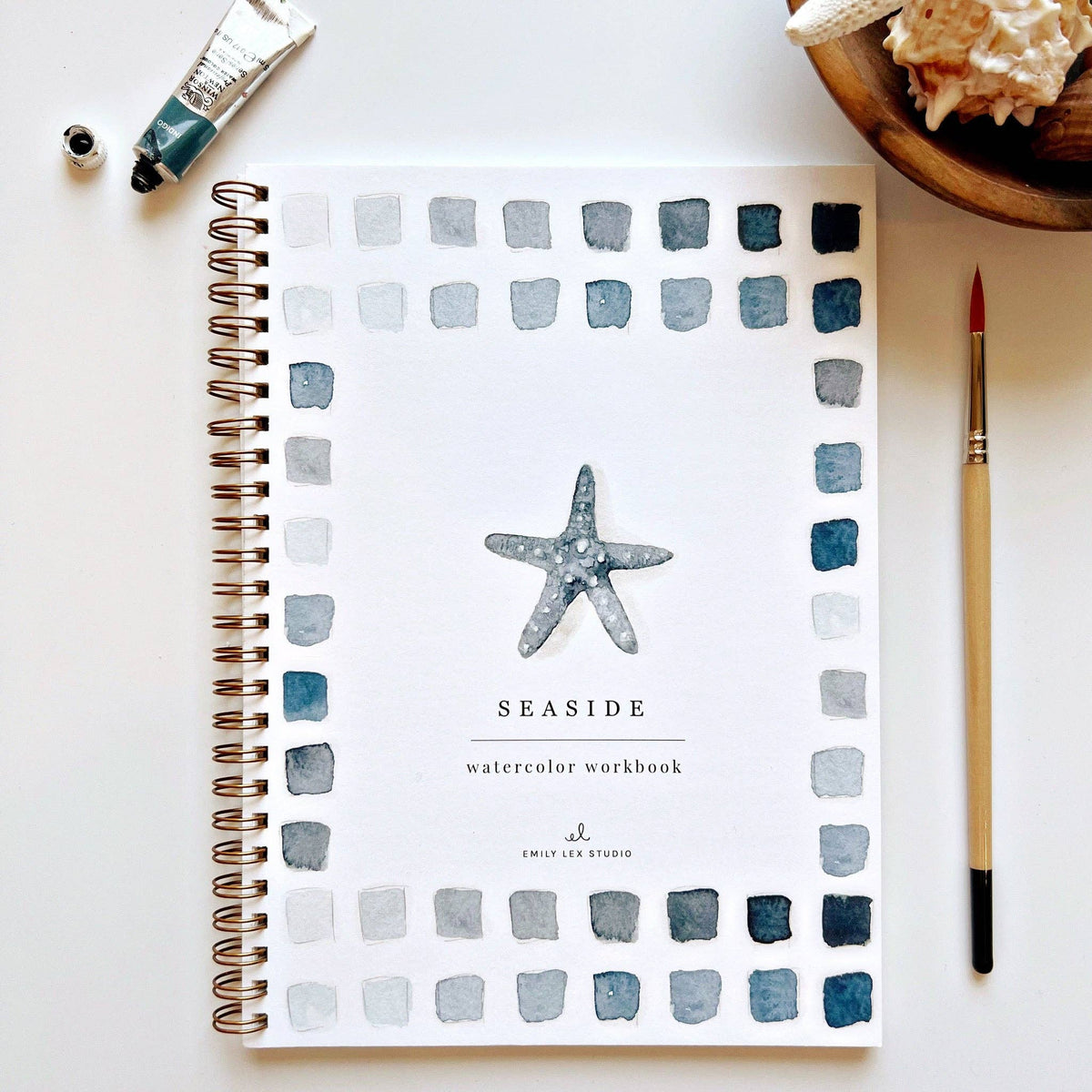 Seaside Watercolor Workbook by Emily Lex Studio — spiral-bound beginner watercolor painting book with a starfish illustration on the cover, bordered by blue-gray watercolor swatches, with a paintbrush and watercolor tube, seashells in background