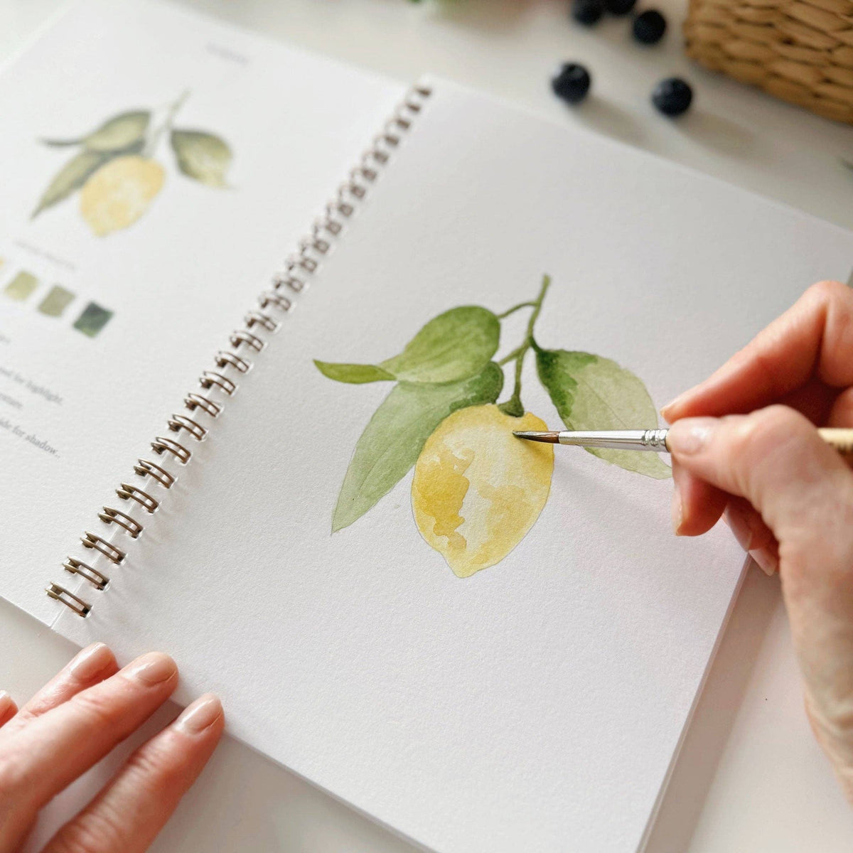 Close-up of hand adding final yellow detail to a nearly complete lemon painting in the Emily Lex Studio Fruit Watercolor Workbook, brush touching the lemon on the sketch page with the completed lemon branch reference illustration visible on the left, blueberries in the backgroun