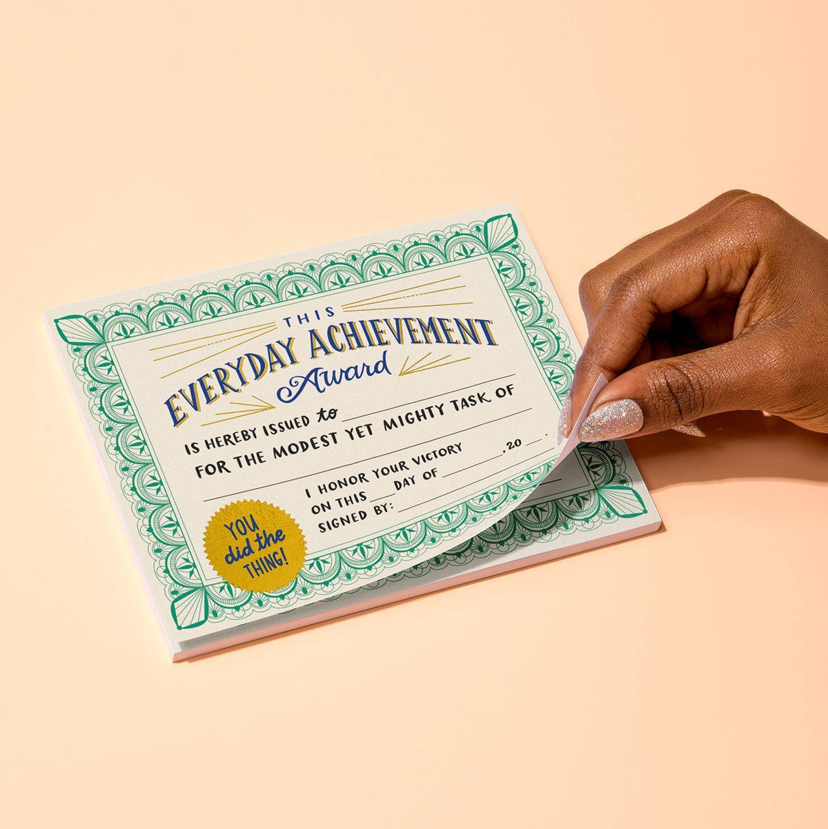 Hand with glittery nails peeling a completed Everyday Achievement Award certificate off the notepad — cream certificate with green border and gold seal on a peach background