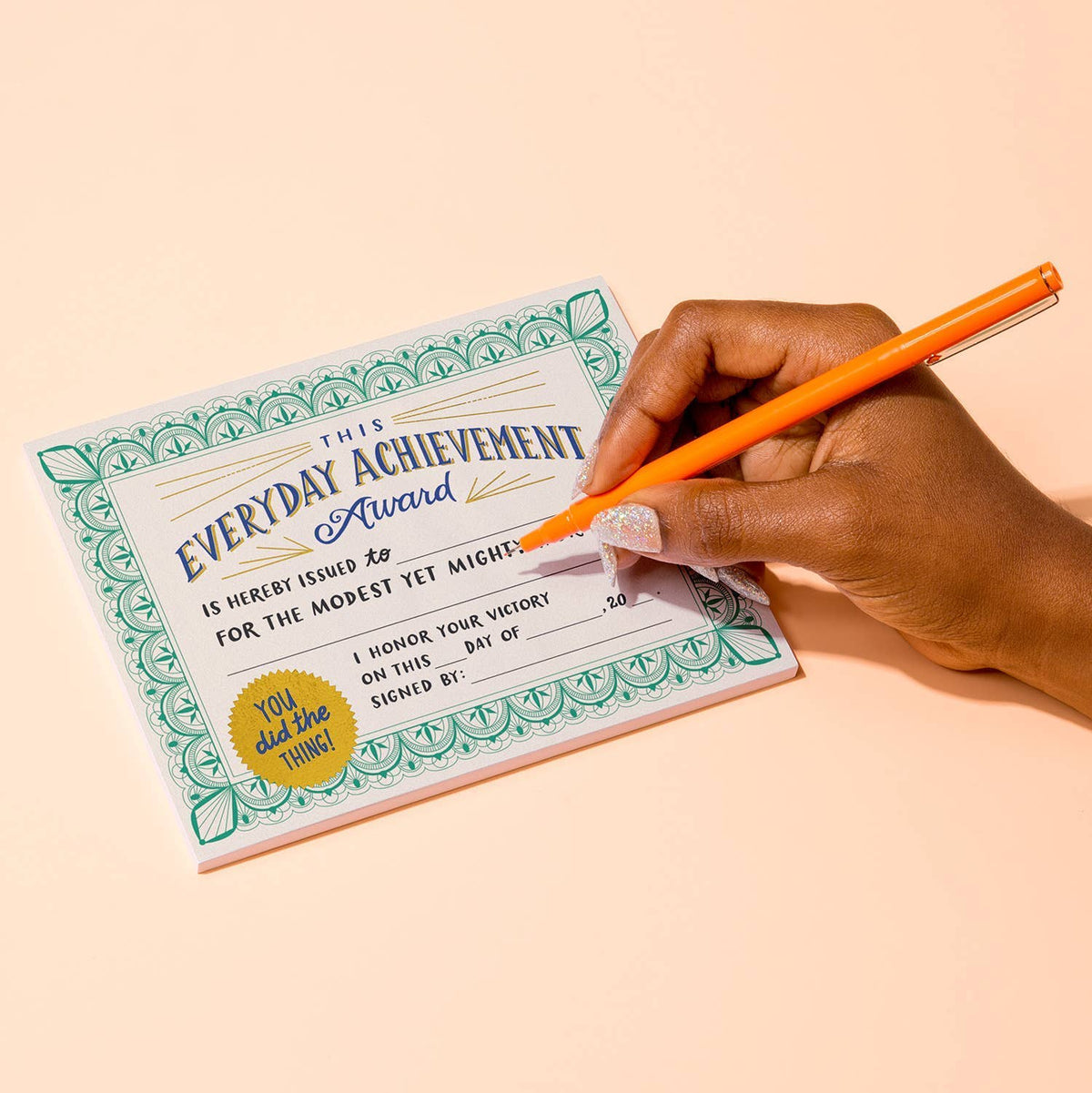 Hand with glittery nails writing on an Everyday Achievement Award certificate with an orange pen — cream certificate with green border and gold seal on a peach background