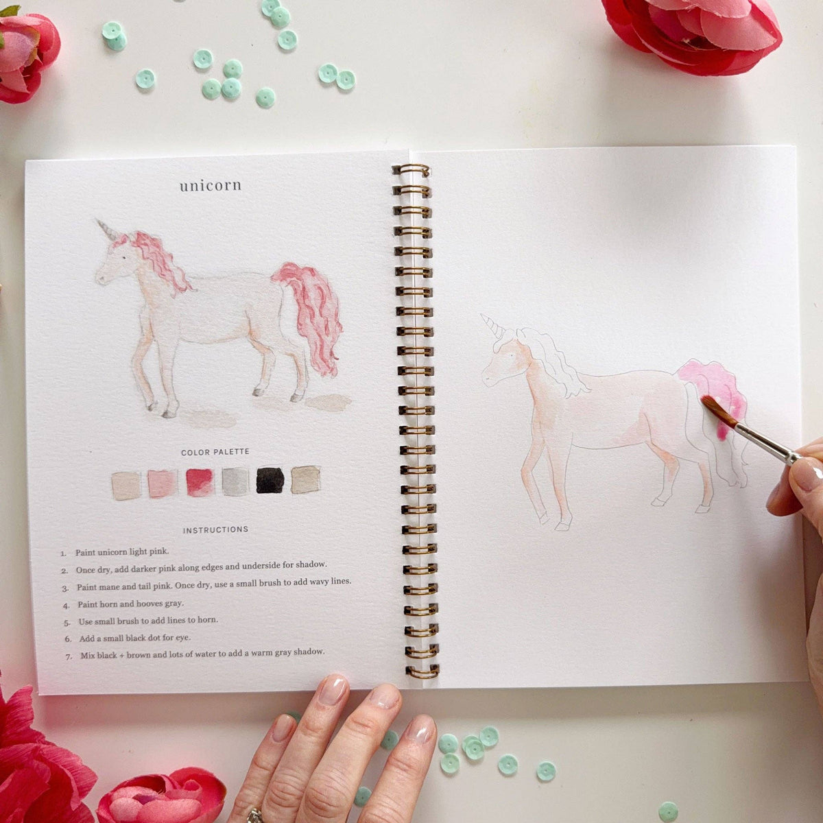 Open Fairytale Watercolor Workbook showing the unicorn lesson; left page has completed watercolor unicorn in pale pink with flowing pink mane and tail, gray horn and hooves, color palette in blush, pink, gray, and black, and 7 instructions; right page shows unicorn sketch being painted in light pink; pink ranunculus and teal sequins in background