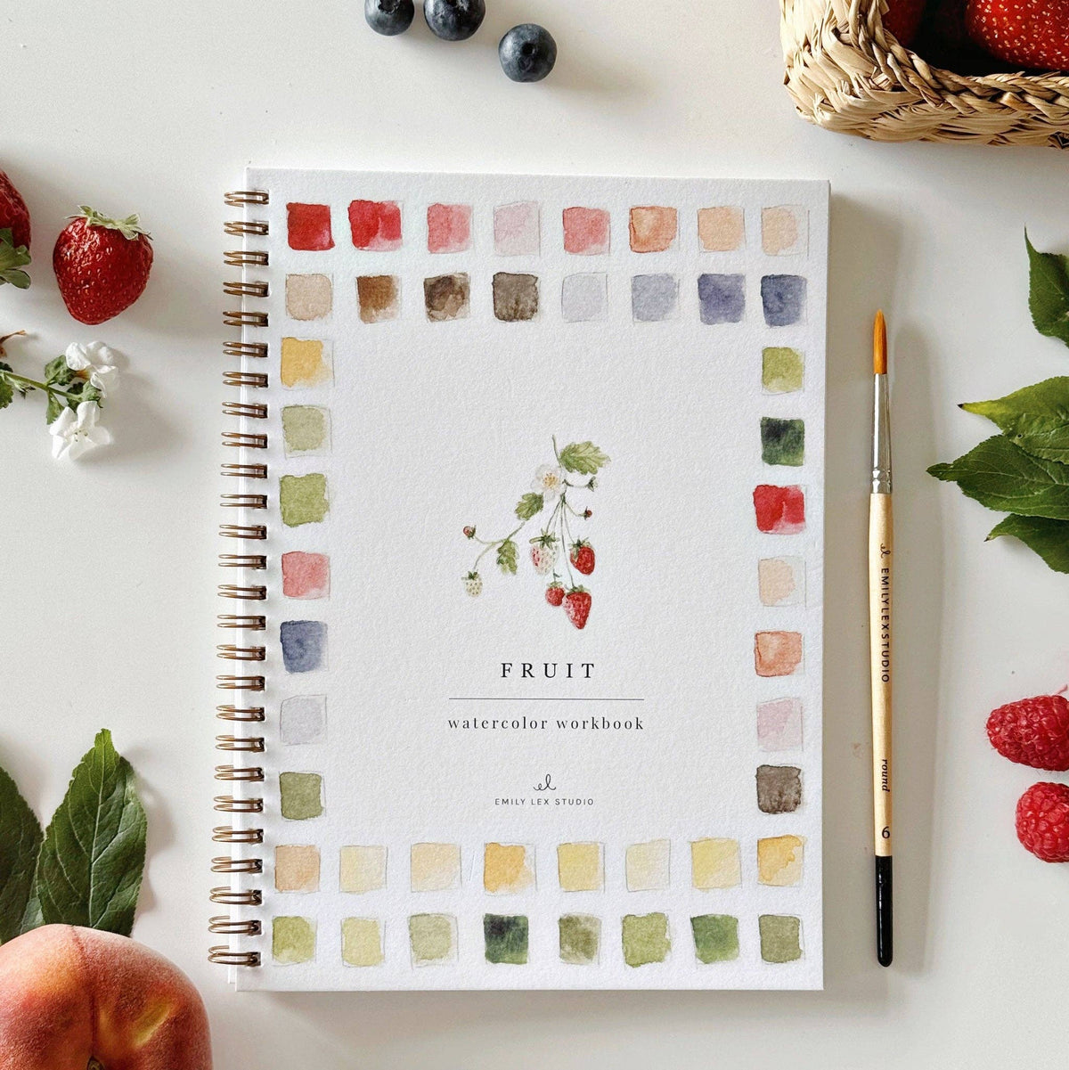 Emily Lex Studio Fruit Watercolor Workbook cover with a strawberry branch illustration surrounded by colorful watercolor paint swatches, styled with a paintbrush beside fresh strawberries, raspberries, blueberries, and mint leaves on a white surface