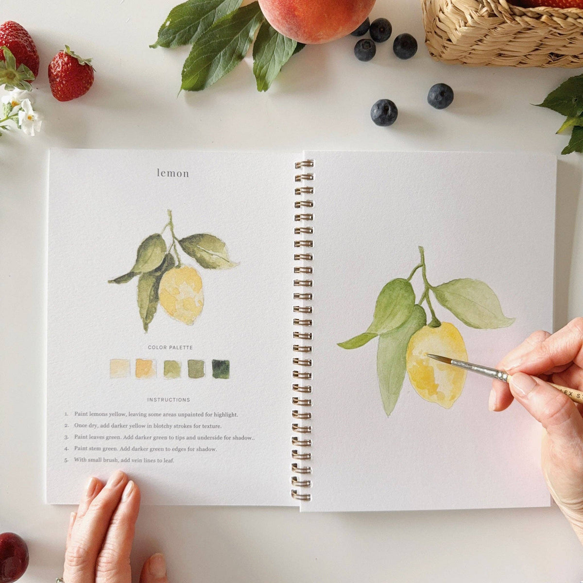 Open spread of Emily Lex Studio Fruit Watercolor Workbook at the lemon lesson — completed yellow lemon branch illustration with color palette and 5-step instructions on the left, hand painting a nearly complete lemon branch on the sketch page on the right