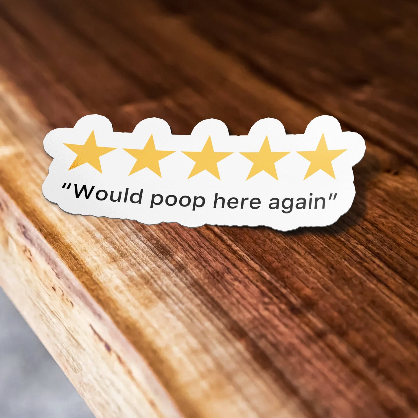 Sticker Bull 5 Stars Would Poop Here Again die cut vinyl sticker laid on a warm wood surface — five golden stars above "Would poop here again" serif quote text with scalloped white border