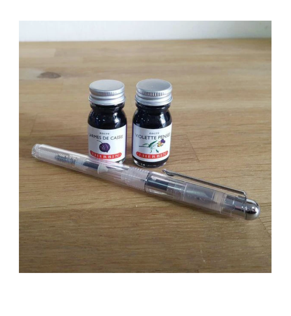 J. Herbin Demonstrator fountain pen laid horizontally on a wood surface next to two small J. Herbin ink bottles — left bottle labeled "Larmes de Cassis" (blackcurrant purple), right bottle labeled "Violette Pensée" (violet); pen shown uncapped with clear barrel and silver trim; bottles have silver metal screw caps and Herbin illustrated labels