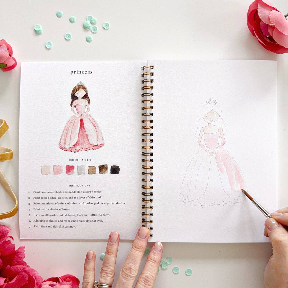Open Fairytale Watercolor Workbook showing the princess lesson; left page has completed watercolor princess in a pink ballgown with tiara, color palette in blush, pink, gray, tan, brown, and black, and 7 instructions; right page shows princess sketch being painted in light pink with fine-tip brush; pink ranunculus and teal sequins in background
