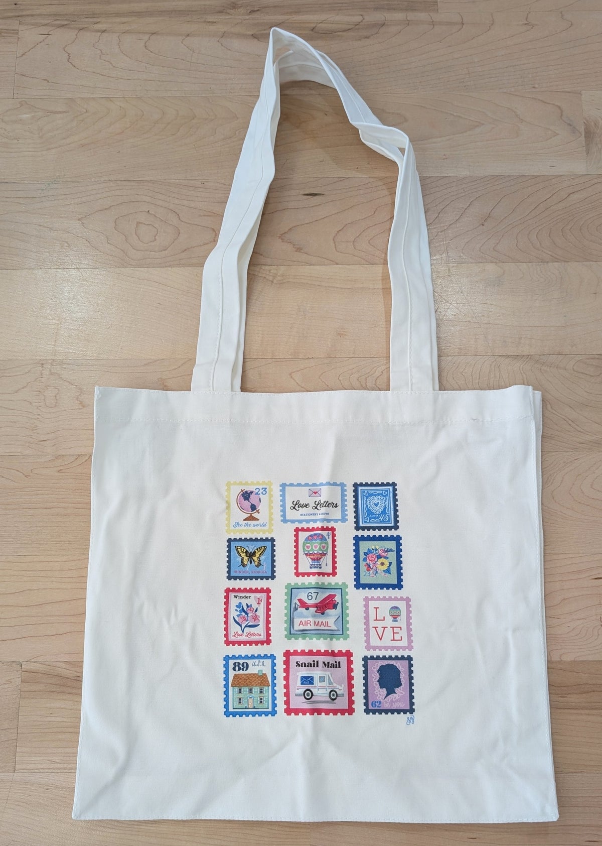 Made by Shellie Love Letters Stamps Canvas Tote — natural canvas tote bag with 12-stamp grid of colorful postage stamp illustrations including globe, Love Letters stamp, butterfly, hot air balloon, floral bouquet, Air Mail biplane, LOVE stamp, house, Snail Mail truck, and Victorian silhouette; flat lay on wood surface