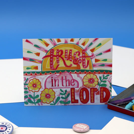 Made by Shellie Trust in the Lord Art Print propped upright on bright blue background — vibrant folk-art watercolor with hand-lettered "Trust in the Lord," golden sunrise with pink and green rays, and yellow flowers; styled with Winder Georgia button, Love Letters pin, and open tin of pastels