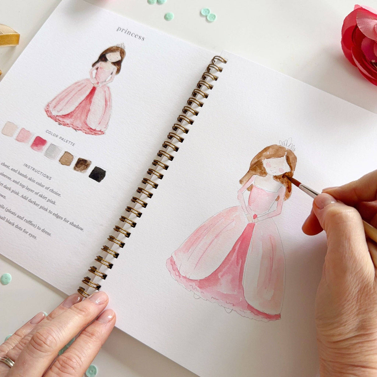 Close-up angled view of the princess lesson; left page shows completed princess and color palette partially visible; right page shows princess sketch nearly complete with pink gown and brown hair being painted by a hand holding a fine-tip brush; pink ranunculus at upper right