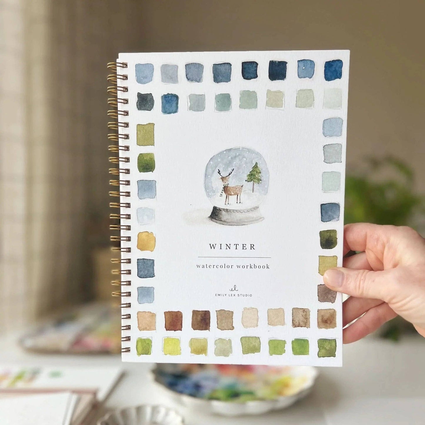 Hand holding the Winter Watercolor Workbook by Emily Lex Studio — cover showing a snow globe with a reindeer and Christmas tree, bordered by a full grid of blue, green, olive, and earth-tone watercolor swatches, with Emily Lex Studio branding