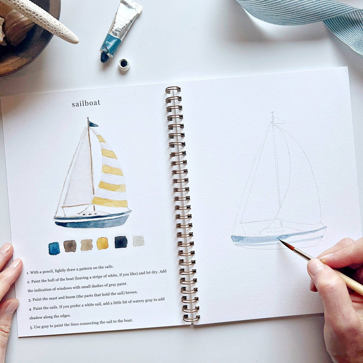 Open spread of Seaside Watercolor Workbook open to sailboat page, hand beginning to paint light blue watercolor on the hull of a sailboat sketch, with completed sailboat illustration, color palette, and step-by-step instructions on left, watercolor tubes and blue ribbon in background