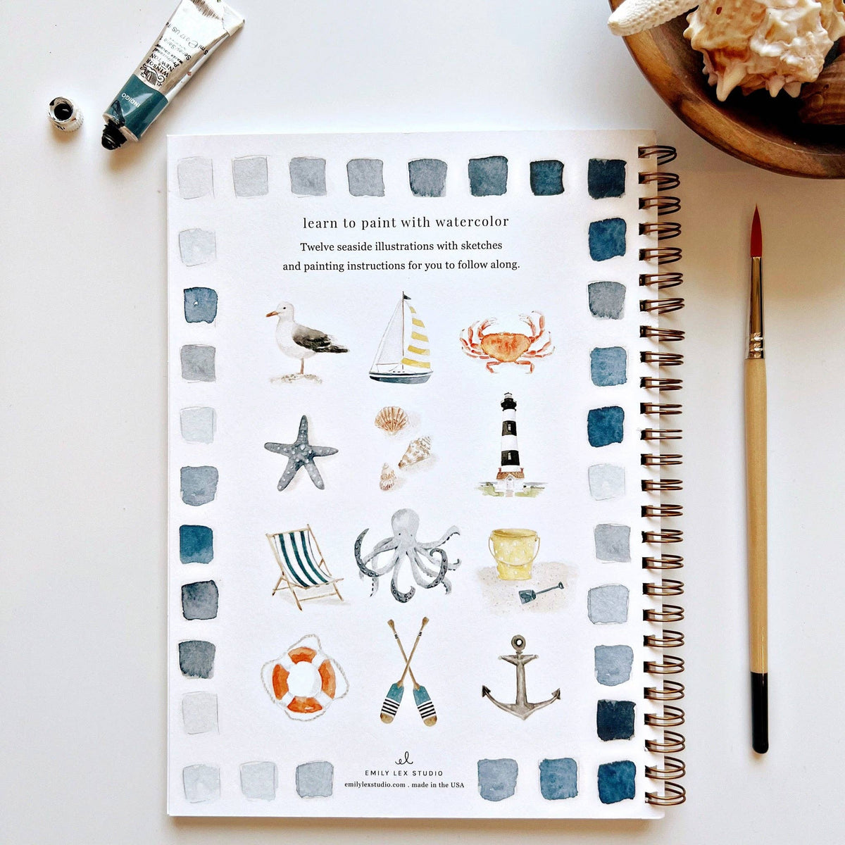 Back cover of Seaside Watercolor Workbook showing all 12 illustrations: seagull, sailboat, crab, starfish, seashells, lighthouse, beach chair, octopus, sand bucket, life ring, oars, and anchor — with blue-gray watercolor swatches border and Emily Lex Studio branding