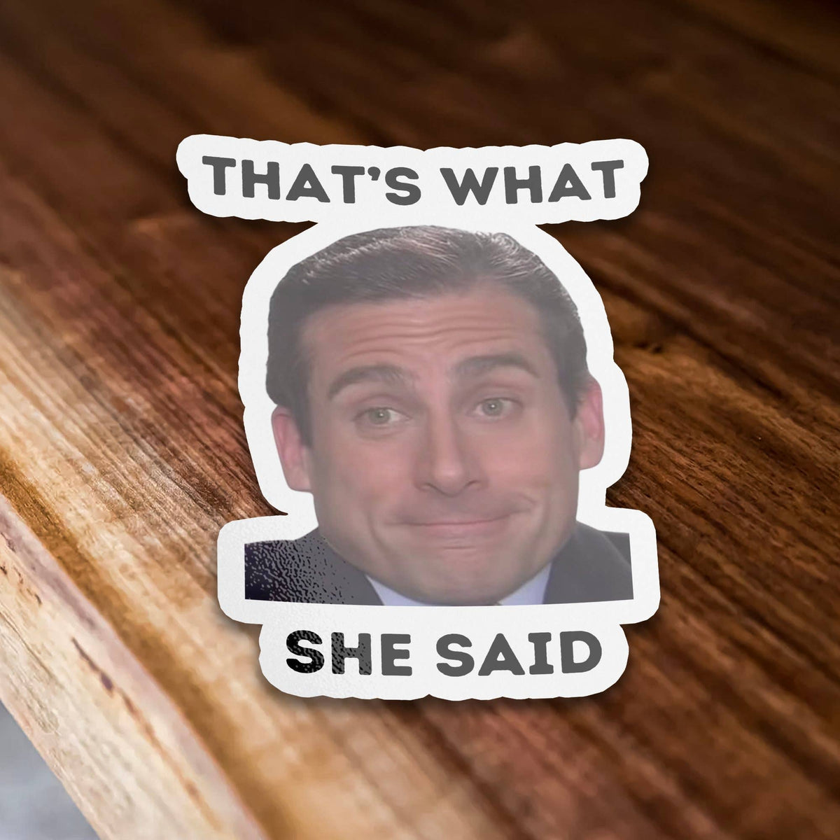 Sticker Bull That's What She Said die cut vinyl sticker propped on a warm wood table edge — photo-real Michael Scott smiling with bold black "THAT'S WHAT / SHE SAID" text; scalloped white die cut border