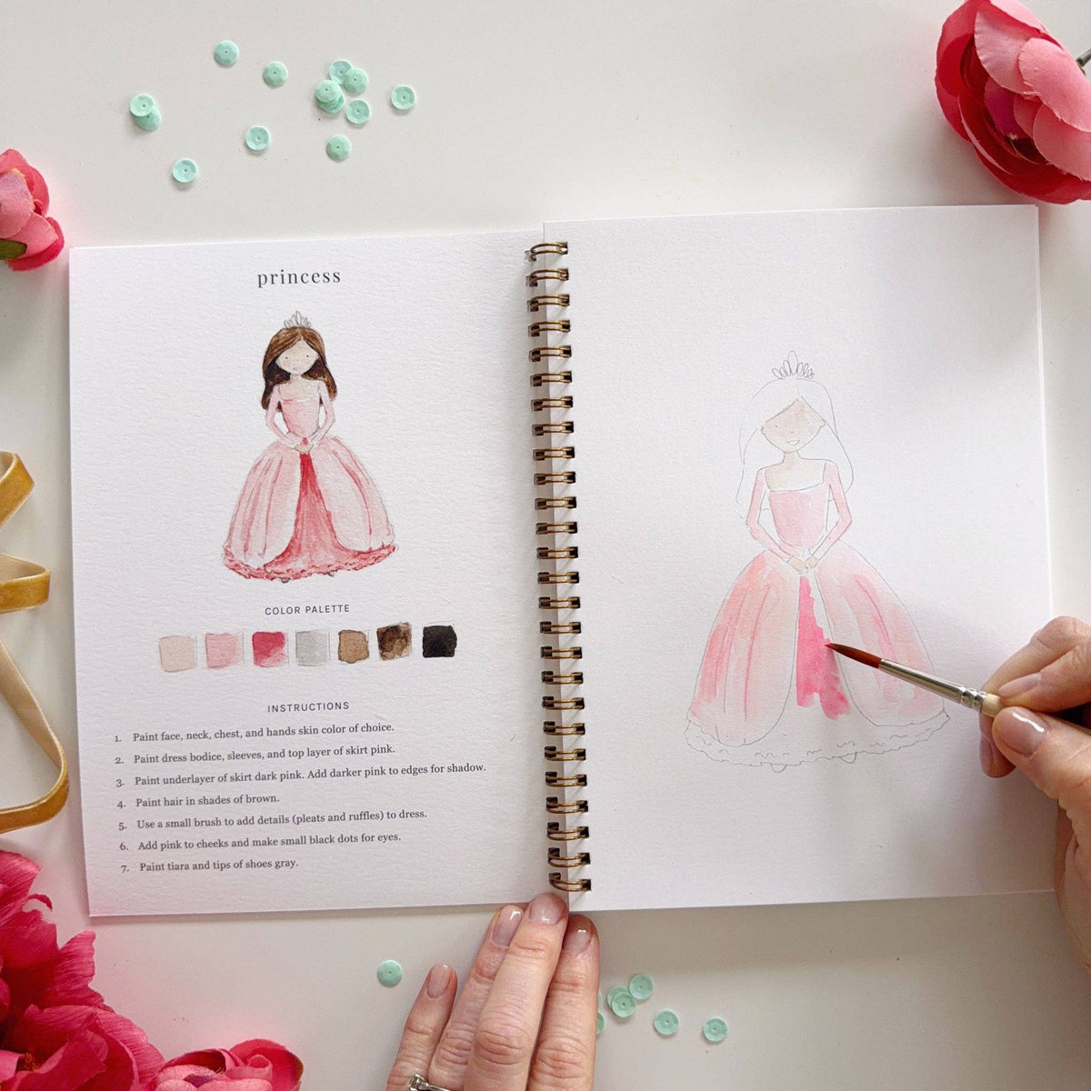 Same princess lesson from a wider angle; left page shows completed princess and color palette; right page shows princess sketch further along with pink skirt painted; two hands visible; pink ranunculus and teal sequins in background