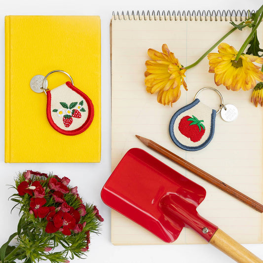 Two Three Potato Four patch keychains styled in a flat lay on a white surface with a yellow hardcover book, cream spiral notebook, orange gerbera daisies, red carnations, two pencils, and a small red enamel garden shovel — left: Strawberry Blossom keychain on cream patch with red border resting on the yellow book; right: Tomato keychain on cream patch with navy border on the notebook; both with silver split rings and brand charms