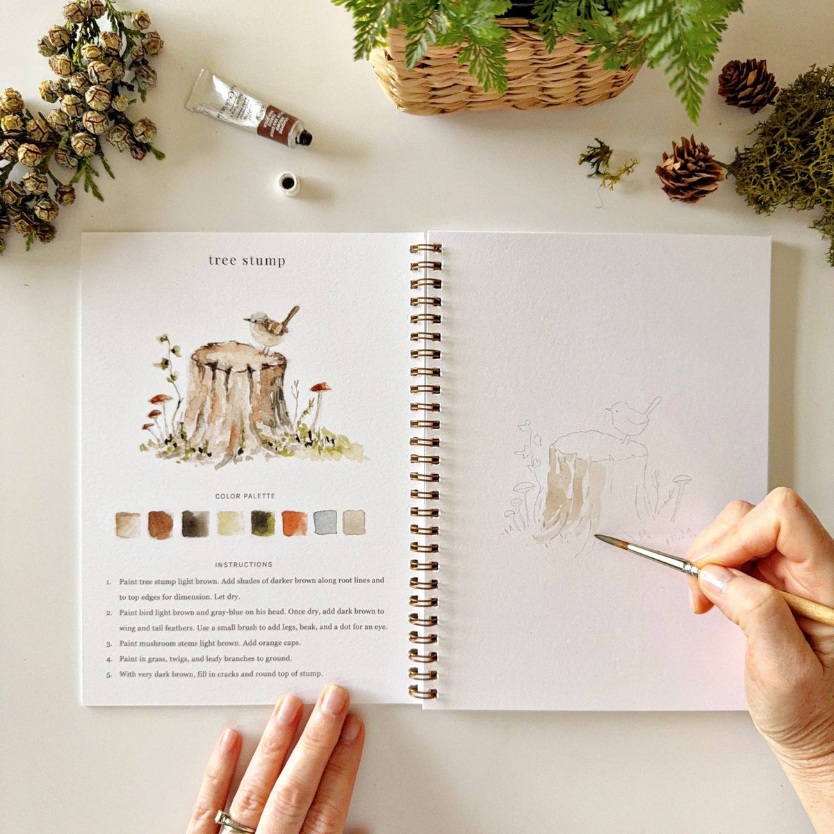 Open spread of Woodland Watercolor Workbook showing tree stump illustration with a wren bird, color palette and instructions on left, hand beginning to paint the tree stump sketch on right