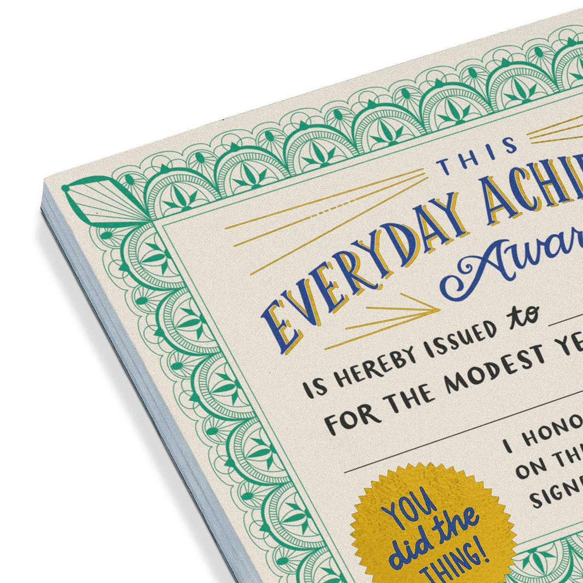 Close-up angled view of the Everyday Achievement Award Certificate Notepad spine — showing the thick stack of 50 cream sheets with green ornate border, blue and gold foil title, and gold seal partially visible