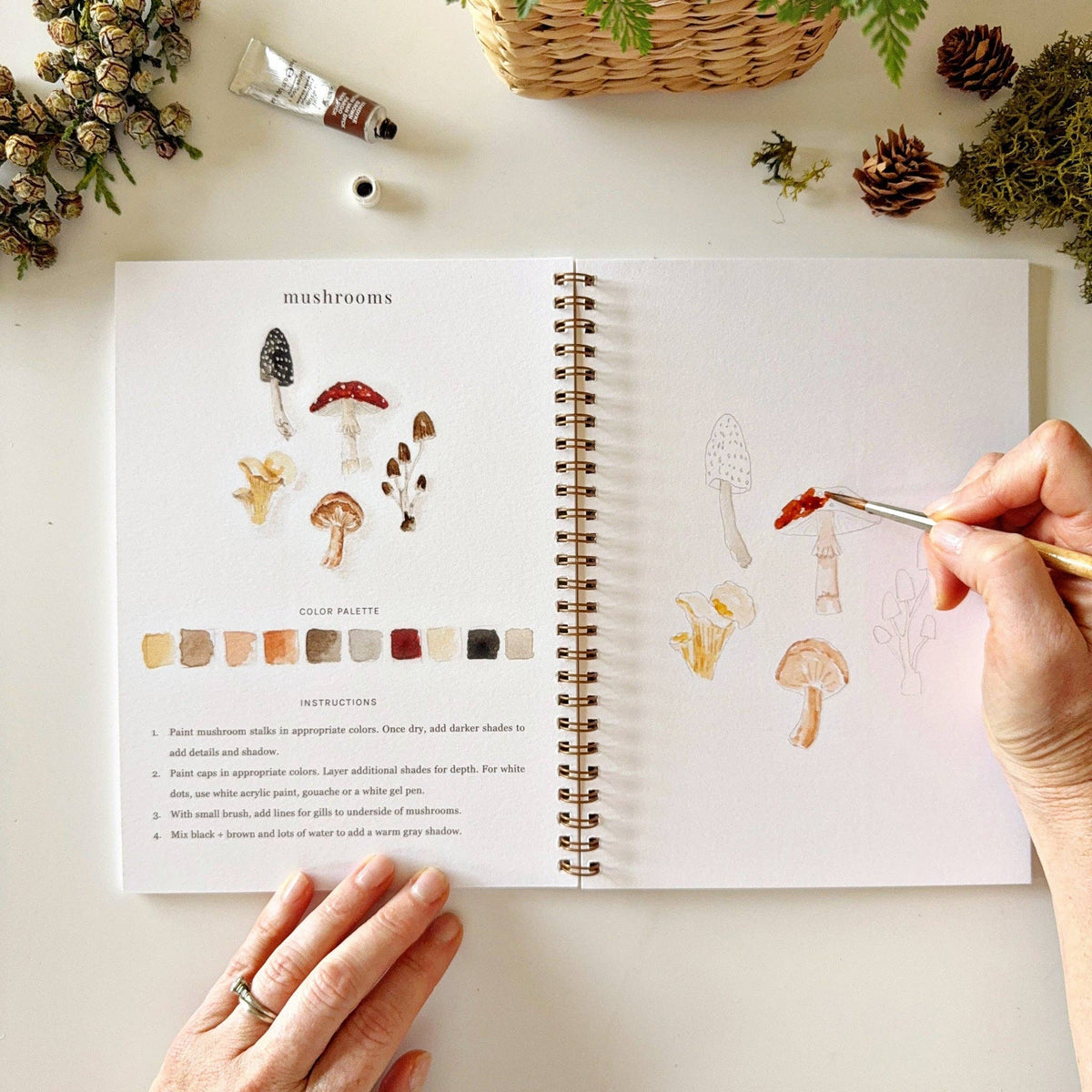 Open spread of Woodland Watercolor Workbook showing mushrooms page, hand painting a red-capped mushroom on the sketch page with color palette and instructions visible on left