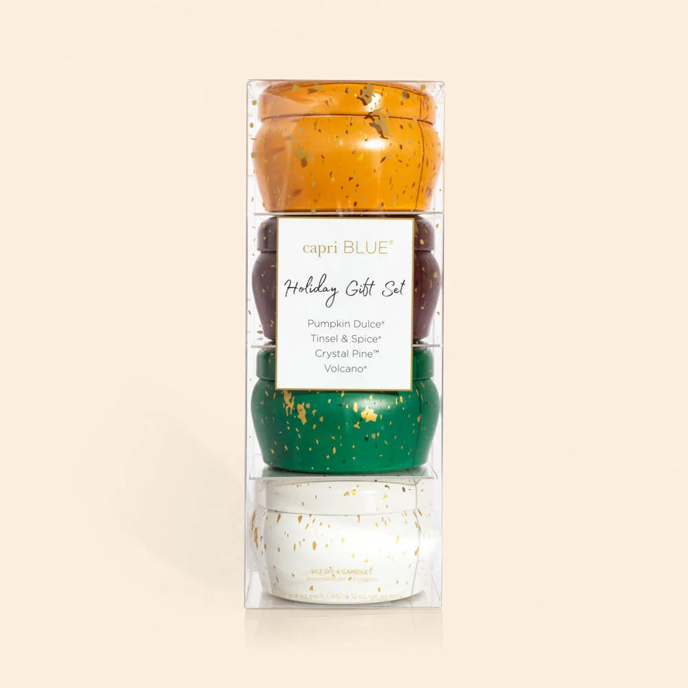 Capri Blue Holiday Gift Set in clear stacking gift box — four gold-splattered mini candle tins stacked vertically in a clear acrylic box; amber Pumpkin Dulce tin at top, burgundy Tinsel & Spice tin second, forest green Crystal Pine tin third, white Volcano tin at bottom; white label at center reading "capri BLUE Holiday Gift Set — Pumpkin Dulce, Tinsel & Spice, Crystal Pine, Volcano"; cream background