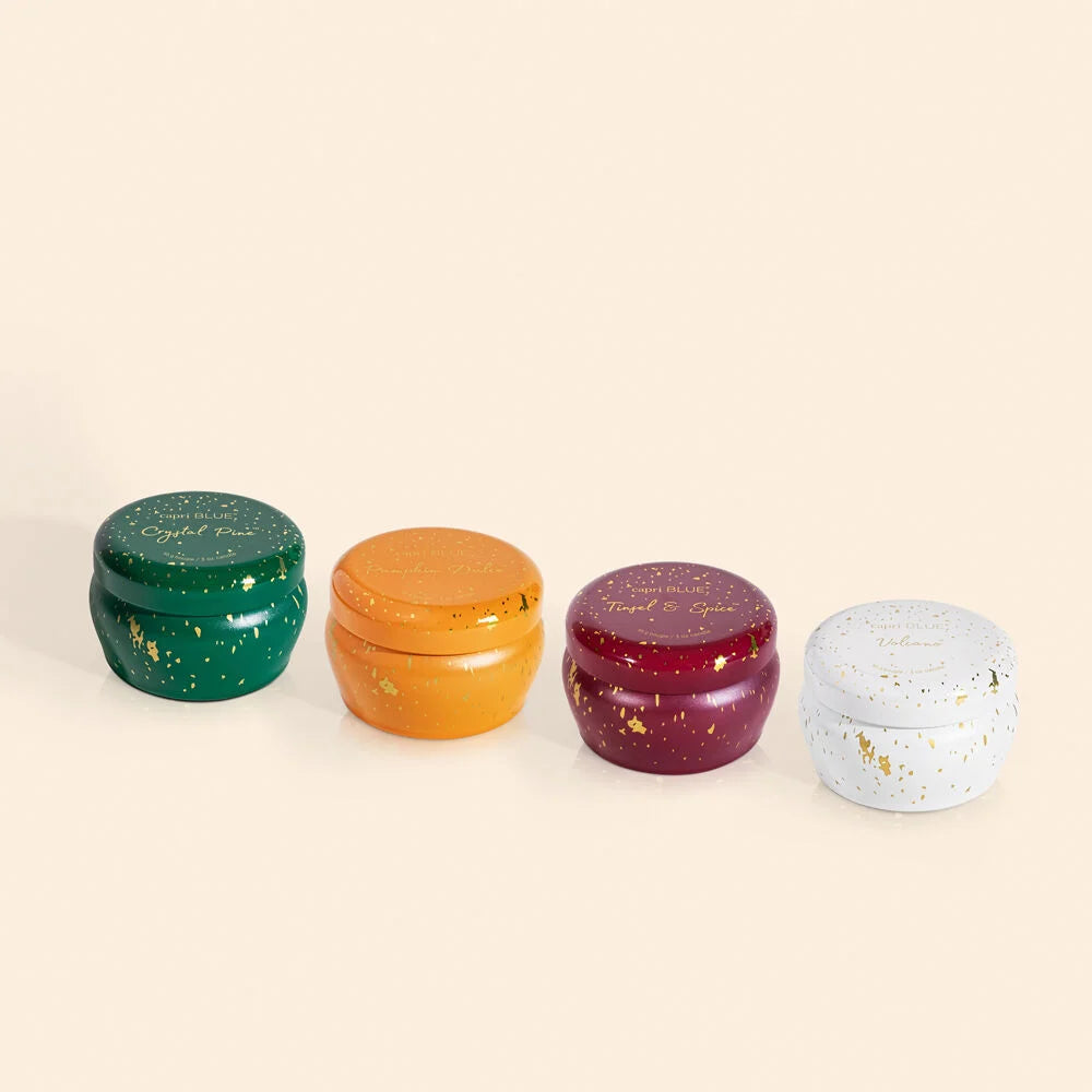 Four Capri Blue mini candle tins in a row — forest green Crystal Pine, amber Pumpkin Dulce, burgundy Tinsel & Spice, and white Volcano tins side by side; all with gold foil splatter finish and Capri Blue script labels on lids; cream background
