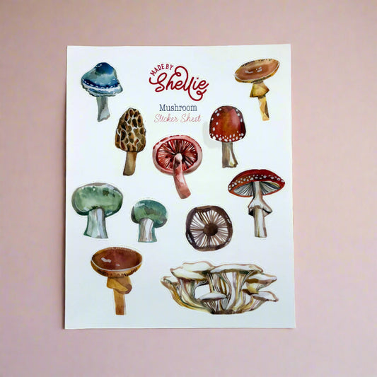 Made by Shellie Mushroom Sticker Sheet — ten watercolor mushroom stickers including blue-capped, morel, fly agaric, red-spotted toadstool, green-capped, brown-gilled, and oyster mushroom cluster; white sheet with Made by Shellie logo; pink background