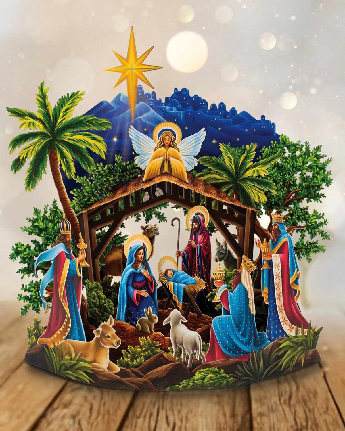 Starlit Nativity pop-up fully open on wood surface — 15-inch 3D nativity scene with Mary in blue kneeling at manger, Joseph with staff, praying angel with gold wings at top, three wise men in colorful robes at left, camel, donkey, lamb, and rabbit at base; lush green palm trees and shrubs; deep navy starlit sky with Bethlehem cityscape and gold Star of Bethlehem; bokeh light background