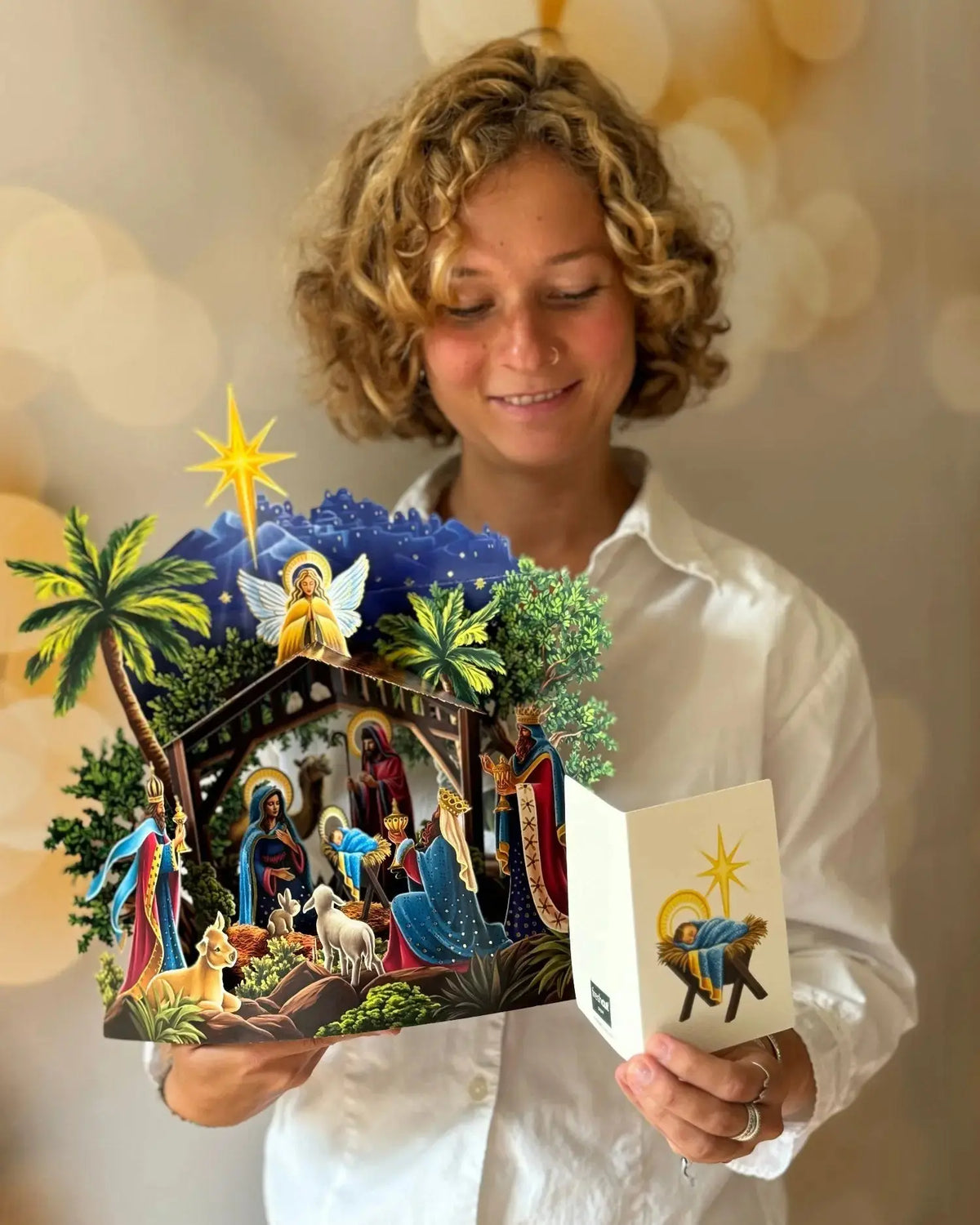 Woman holding open Starlit Nativity pop-up card — smiling woman in white shirt holding fully open 15-inch nativity pop-up in both hands; note card with baby Jesus in manger illustration held alongside; warm bokeh background