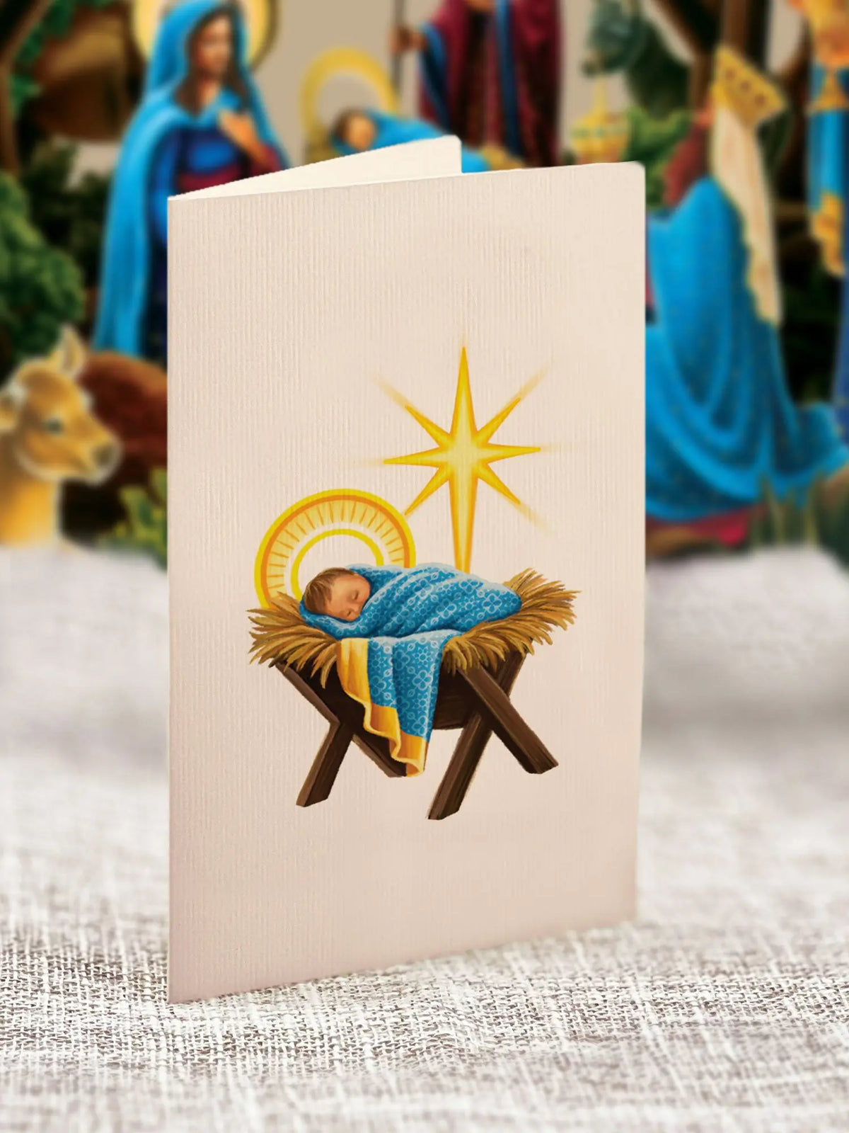 Starlit Nativity note card close-up — cream folded note card with illustration of baby Jesus swaddled in blue blanket in a hay manger with gold halo and gold eight-pointed star; pop-up nativity scene blurred in background