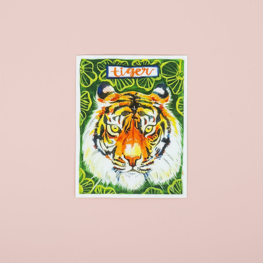 Made by Shellie Tiger vinyl sticker — bold watercolor tiger face with orange and black stripes, golden eyes, and white cheeks on lush green tropical leaf background; "tiger" hand-lettered in blue banner at top; rectangular with white border; pink background