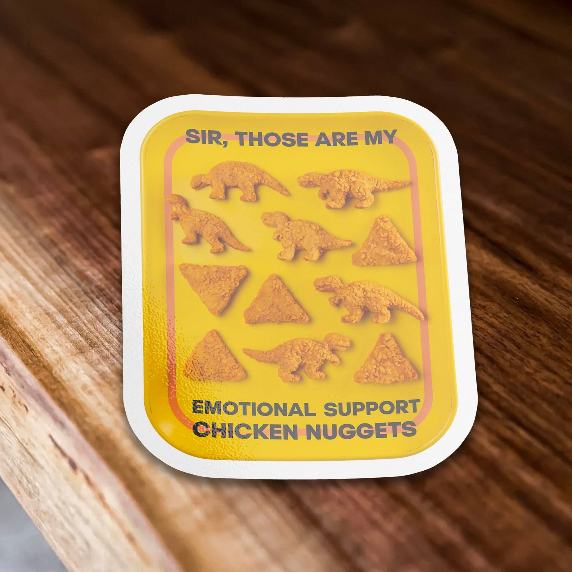 Sticker Bull Sir, Those Are My Emotional Support Chicken Nuggets die cut vinyl sticker laid on a warm wood surface — yellow tray with dino nuggets and bold black text; white die cut border