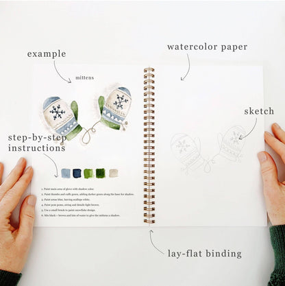 Open spread of Winter Watercolor Workbook open to mittens page, hands holding the book showing completed blue and green fair isle mittens illustration with color palette and step-by-step instructions on left, blank mitten sketch on right with labeled callouts for example, sketch, watercolor paper, step-by-step instructions, and lay-flat binding