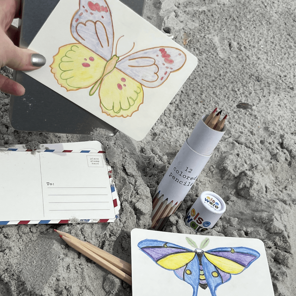 Lifestyle flat lay on sandy beach surface — adult hand with silver nail polish holding a completed Elseware Unplug coloring postcard with a yellow and pink butterfly colored in pencil; classic air-mail address postcard face-down in center; Elseware Unplug 12-colored pencil tube standing upright in sand; second completed postcard at bottom showing a blue and yellow luna moth colored with pencils; two loose colored pencils beside