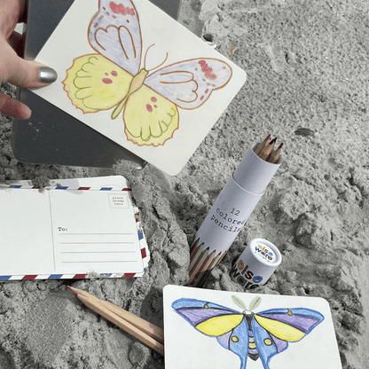 Lifestyle flat lay on sandy beach surface — adult hand with silver nail polish holding a completed Elseware Unplug coloring postcard with a yellow and pink butterfly colored in pencil; classic air-mail address postcard face-down in center; Elseware Unplug 12-colored pencil tube standing upright in sand; second completed postcard at bottom showing a blue and yellow luna moth colored with pencils; two loose colored pencils beside