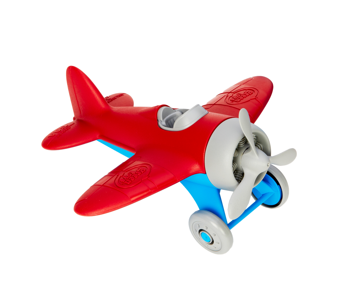 Green Toys Airplane in red and blue on white background — chunky red plastic single-seater propeller plane with rounded red wings, red fuselage, light blue undercarriage and landing gear struts, two grey rolling wheels, grey three-blade spinning propeller, open grey cockpit, and Green Toys logo embossed on the wings; viewed from front-right angle