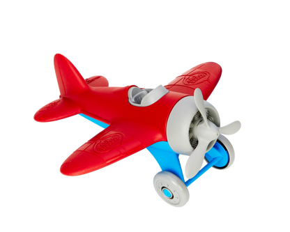 Green Toys Airplane in red and blue on white background — chunky red plastic single-seater propeller plane with rounded red wings, red fuselage, light blue undercarriage and landing gear struts, two grey rolling wheels, grey three-blade spinning propeller, open grey cockpit, and Green Toys logo embossed on the wings; viewed from front-right angle