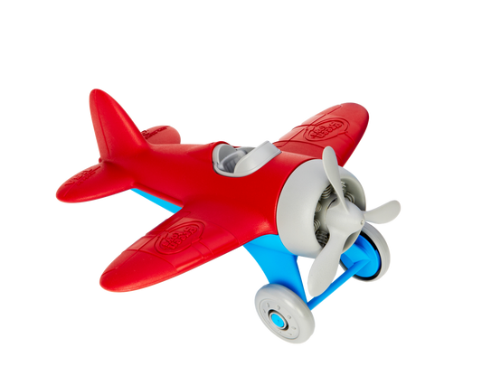 Green Toys Airplane in red and blue on white background — chunky red plastic single-seater propeller plane with rounded red wings, red fuselage, light blue undercarriage and landing gear struts, two grey rolling wheels, grey three-blade spinning propeller, open grey cockpit, and Green Toys logo embossed on the wings; viewed from front-right angle