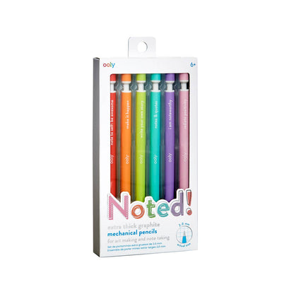 OOLY Noted! Extra Thick Graphite Mechanical Pencils set of 6 in retail packaging on white background — side-angle three-quarter view of the box showing the grey side panel and front clear window; six rainbow pencils visible inside in red, orange, lime, teal, purple, and pink order; "Noted!" multicolor logo, "extra thick graphite mechanical pencils" subtitle, 2.0mm tip diagram, and ooly logo with 6+ age label at top visible on the front face