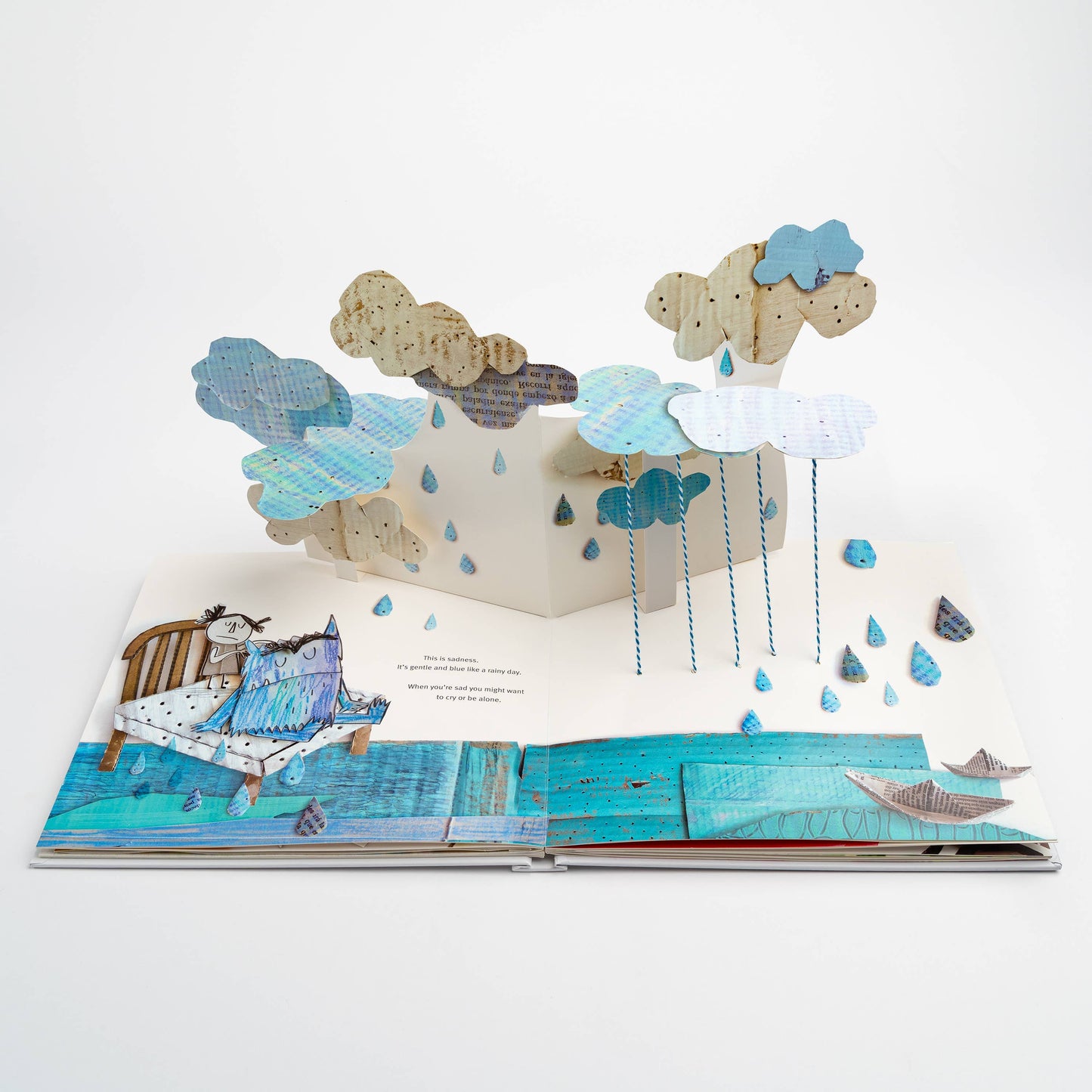 Open spread of The Color Monster: A Pop-Up Book of Feelings showing the sadness page — elaborate 3-D pop-up of layered blue and cream rain clouds with hanging blue raindrop strings above a blue watercolor floor with paper boats, and the blue monster sitting on a bed with the girl, text reading "This is sadness. It's gentle and blue like a rainy day."