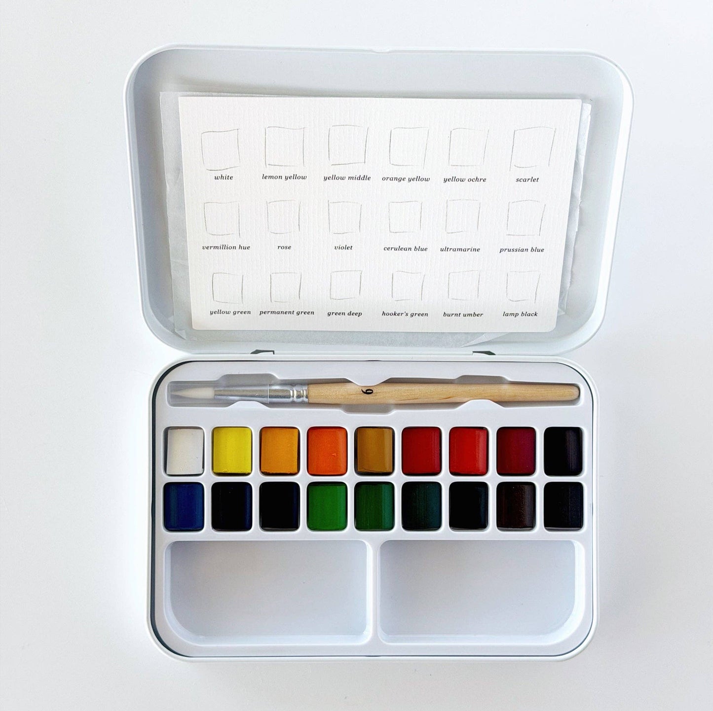 Emily Lex Studio watercolor paint set open flat on white surface; tin lid shows color swatch card with 18 labeled colors: white, lemon yellow, yellow middle, orange yellow, yellow ochre, scarlet, vermillion hue, rose, violet, cerulean blue, ultramarine, prussian blue, yellow green, permanent green, green deep, hooker's green, burnt umber, and lamp black; tin base holds 18 paint pans in two rows with two mixing trays below and a size 6 round brush resting across the top